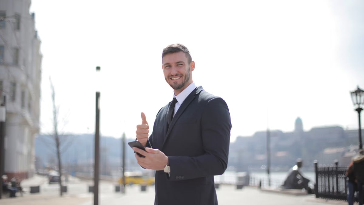 Smiling businessman in a suit giving thumbs up and holding a smartphone outdoors in a city setting.